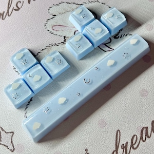 May include: Light blue keyboard keycaps with cloud and star designs. The keycaps are arranged in two groups, with a long space bar keycap. The keycaps have a glossy finish and are set against a pink background with cursive text.