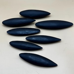 May include: Seven smooth, dark blue, elongated, oval-shaped wooden pieces. The pieces are arranged in a semi-circle on a light beige surface.