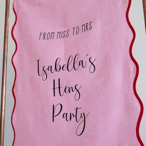 K&ouml;nnte beinhalten: Rosa Stoffbanner mit rotem, gewelltem Rand, das an einem Kupferrohr h&auml;ngt. Das Banner lautet "From Miss to Mrs Isabella's Hens Party 22.09.2023 35 Days to Go".