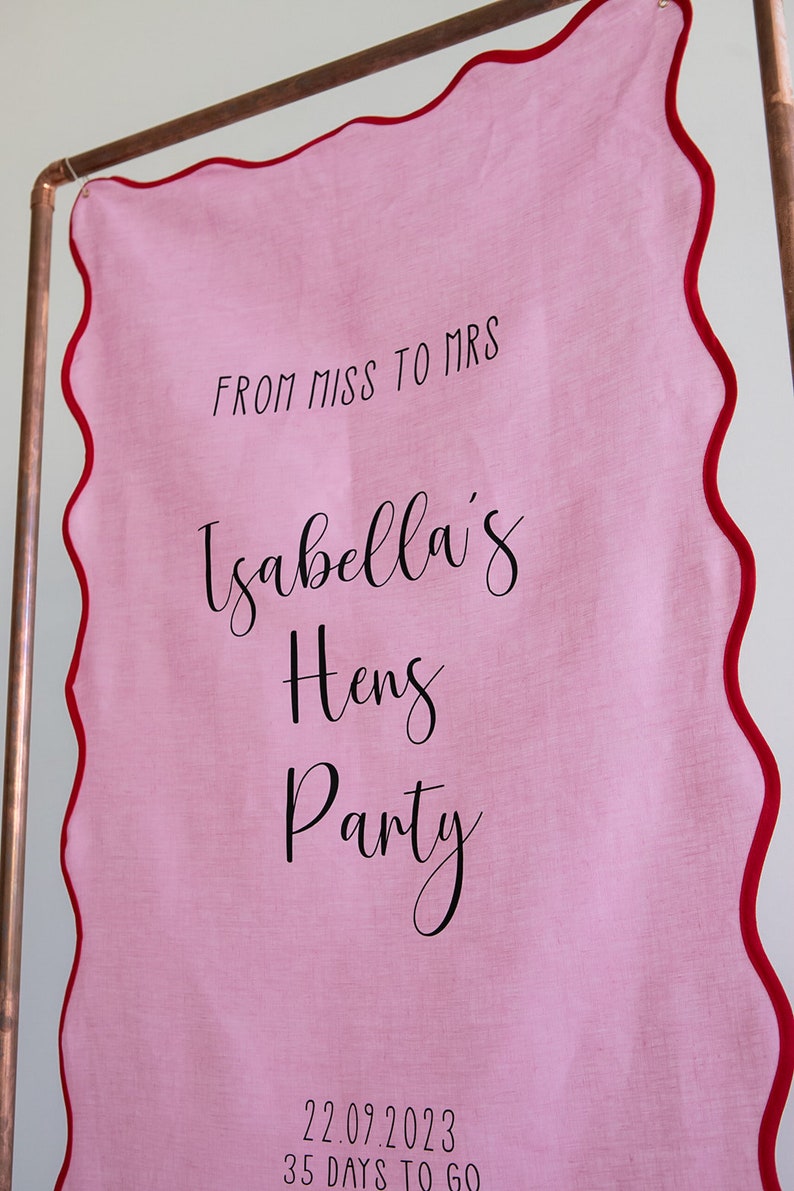 K&ouml;nnte beinhalten: Rosa Stoffbanner mit rotem, gewelltem Rand, das an einem Kupferrohr h&auml;ngt. Das Banner lautet "From Miss to Mrs Isabella's Hens Party 22.09.2023 35 Days to Go".