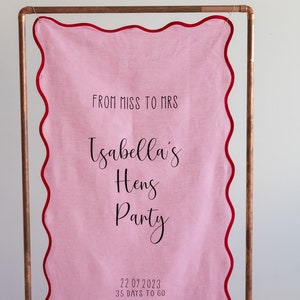K&ouml;nnte beinhalten: Rosa Stoffbanner mit gewellten R&auml;ndern und rotem Besatz, das an einem Kupferrohr h&auml;ngt. Das Banner lautet "From Miss to Mrs Isabella's Hens Party 22.07.2023 35 Days to Go".