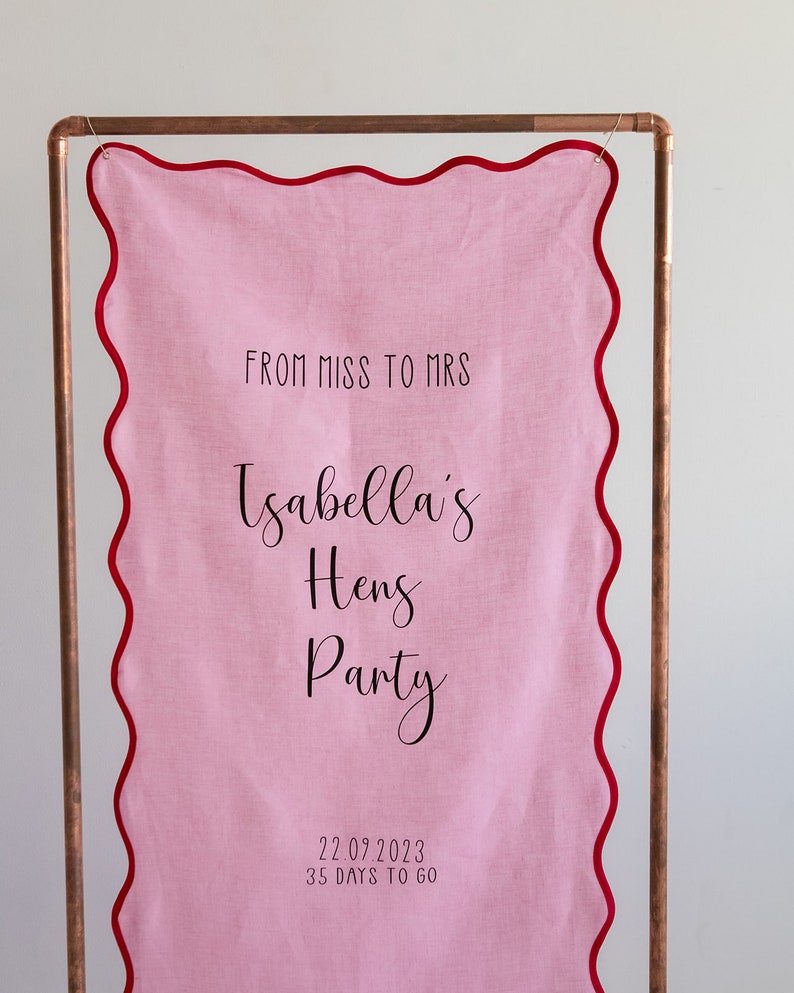 K&ouml;nnte beinhalten: Rosa Stoffbanner mit gewellten R&auml;ndern und rotem Besatz, das an einem Kupferrohr h&auml;ngt. Das Banner lautet "From Miss to Mrs Isabella's Hens Party 22.07.2023 35 Days to Go".