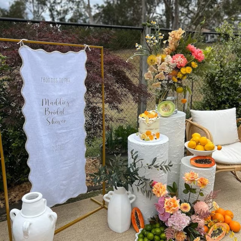 K&ouml;nnte beinhalten: Ein Brautdusche-Setup mit einem wei&szlig;en Schild, auf dem "Von Miss zu Mrs. Maddies Brautdusche" steht. Das Schild steht auf einem goldenen St&auml;nder. Es gibt wei&szlig;e S&auml;ulen mit einem Kuchen und Obst darauf. Au&szlig;erdem gibt es wei&szlig;e Vasen mit Blumen und Gr&uuml;npflanzen.