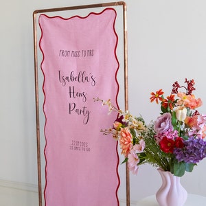 K&ouml;nnte beinhalten: Rosa Stoffbanner mit dem Text "From Miss to Mrs Isabella's Hens Party 22.07.2023 35 Days to Go" an einem Kupferrohrrahmen. Das Banner hat einen gewellten Rand und ist mit einem bunten Blumenstrau&szlig; verziert.