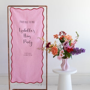 K&ouml;nnte beinhalten: Ein rosa Stoffbanner mit gewelltem Rand h&auml;ngt an einem Kupferrohrrahmen. Das Banner lautet "From Miss to Mrs. Isabella's Hens Party 22.07.2023 35 Days to Go". Ein wei&szlig;er Podesttisch mit einer rosa Vase gef&uuml;llt mit bunten Blumen steht vor dem Banner.