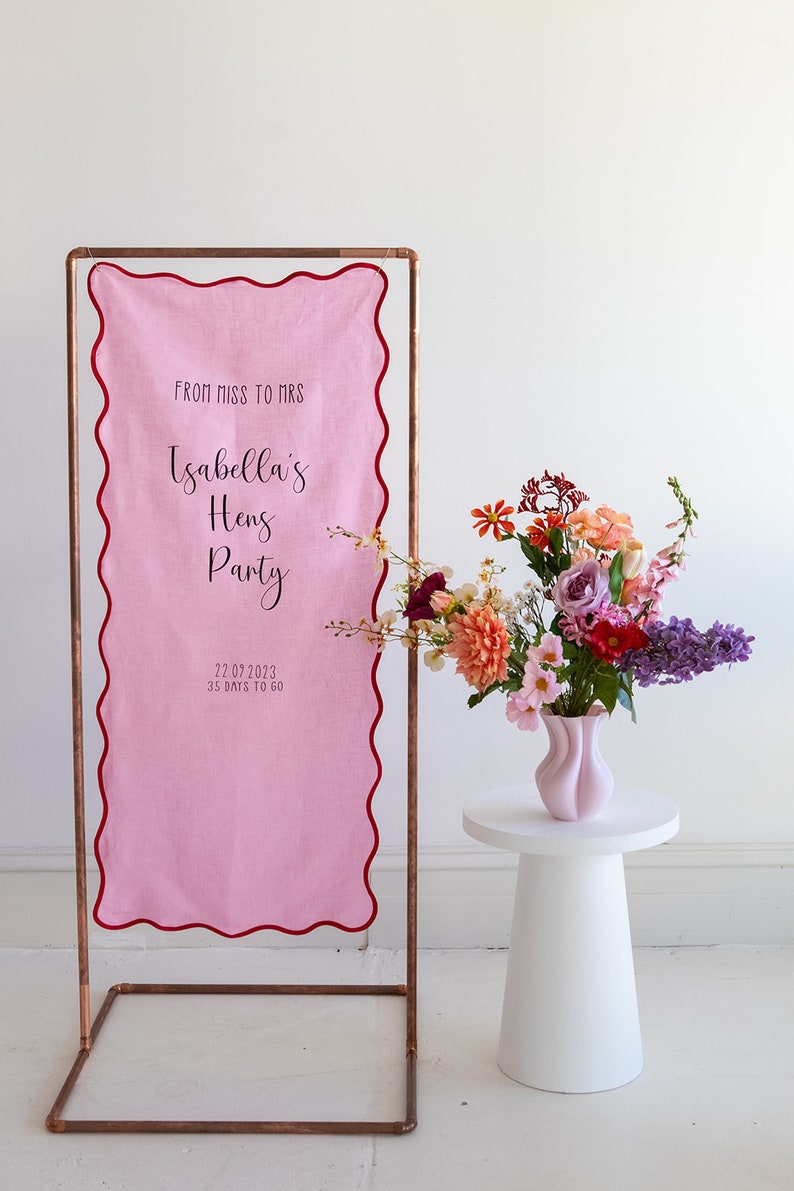 K&ouml;nnte beinhalten: Ein rosa Stoffbanner mit gewelltem Rand h&auml;ngt an einem Kupferrohrrahmen. Das Banner lautet "From Miss to Mrs. Isabella's Hens Party 22.07.2023 35 Days to Go". Ein wei&szlig;er Podesttisch mit einer rosa Vase gef&uuml;llt mit bunten Blumen steht vor dem Banner.