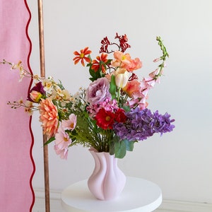 K&ouml;nnte beinhalten: Eine rosa Keramikvase mit einem einzigartigen, wellenf&ouml;rmigen Design h&auml;lt einen bunten Blumenstrau&szlig;. Der Strau&szlig; enth&auml;lt rosa, lila, rote, orangefarbene und gelbe Blumen. Die Vase steht auf einem wei&szlig;en Podesttisch.