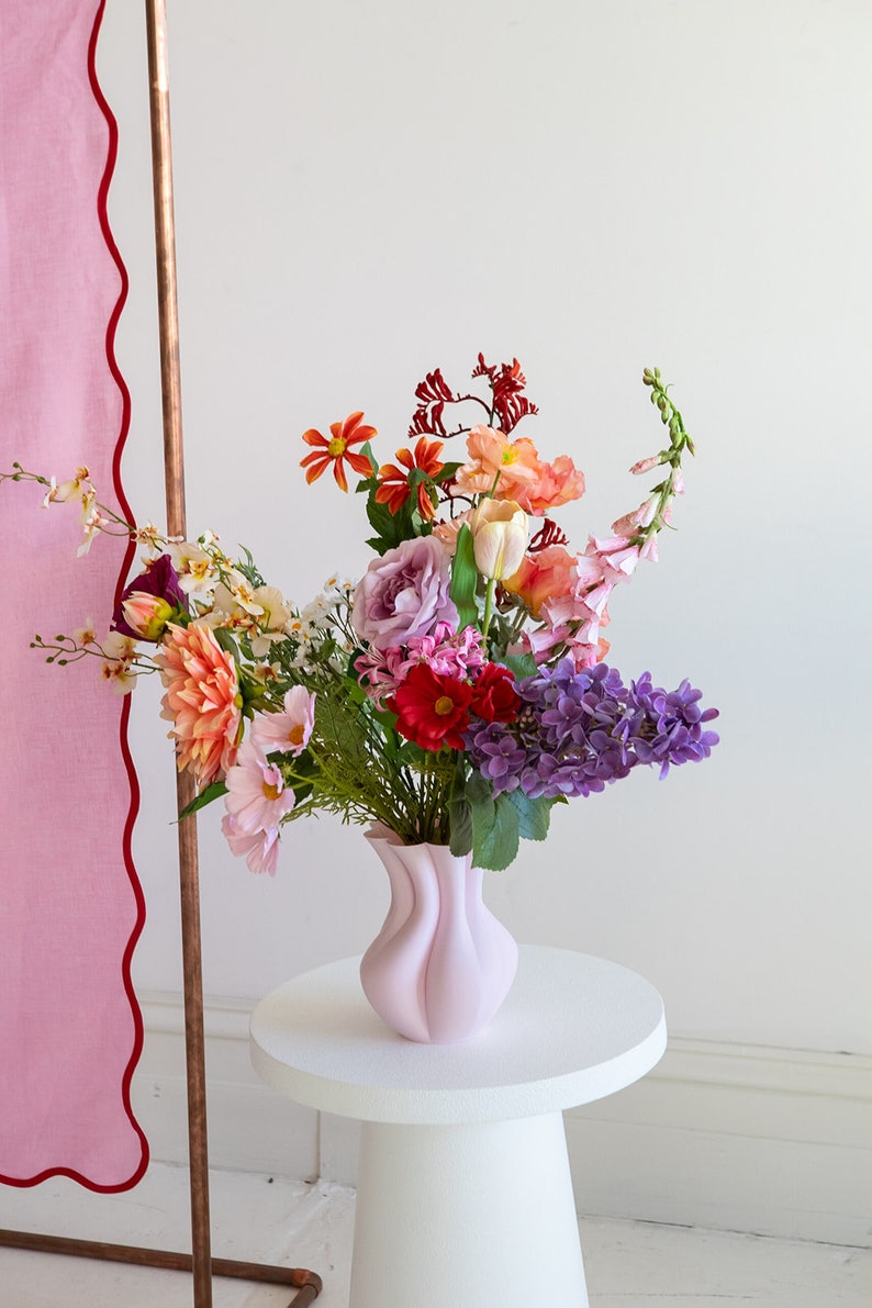 K&ouml;nnte beinhalten: Eine rosa Keramikvase mit einem einzigartigen, wellenf&ouml;rmigen Design h&auml;lt einen bunten Blumenstrau&szlig;. Der Strau&szlig; enth&auml;lt rosa, lila, rote, orangefarbene und gelbe Blumen. Die Vase steht auf einem wei&szlig;en Podesttisch.