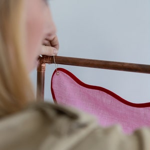 K&ouml;nnte beinhalten: Eine Person h&auml;ngt ein rosafarbenes Stoffbanner mit rotem Rand an einem Kupferrohr. Das Banner hat einen wellenf&ouml;rmigen Rand und ist mit einem Metallring befestigt.