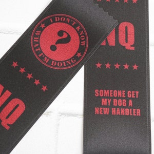 May include: Two black satin ribbons with red text. The top ribbon says "I don't know what I'm doing" with a question mark in a circle. The bottom ribbon says "NQ". The second ribbon says "NQ" and "Someone get my dog a new handler".
