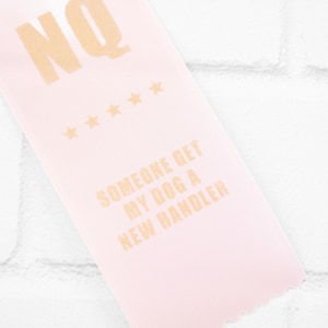 May include: Pink ribbon with gold text that reads "NQ" and "Someone get my dog a new handler". There are five gold stars above the text "NQ".