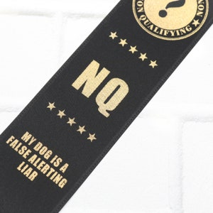 May include: Black ribbon with gold text that reads "NQ" and "My dog is a false alerting liar". The ribbon also has five gold stars and a gold circle with a question mark and the text "Non Qualifying".