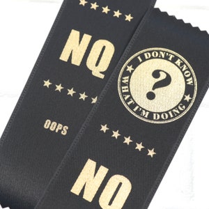 May include: Two black satin ribbons with gold text. The first ribbon says "NO" with five gold stars above the word and "OOPS" below. The second ribbon says "I DON'T KNOW WHAT I'M DOING" in a circle with a question mark in the center and five gold stars around the circle. The word "NO" is below the circle.