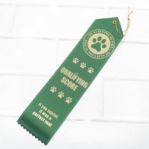 May include: Green ribbon with gold paw prints and the text "Qualifying Score". The ribbon has a gold circle with a paw print and the text "Qualifying Score". The text "If you squint, it was a perfect run!" is printed on the ribbon.