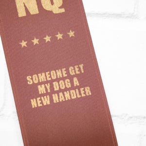 May include: A maroon ribbon with gold text and five gold stars. The text reads "SOMEONE GET MY DOG A NEW HANDLER". The ribbon is angled against a white brick wall.