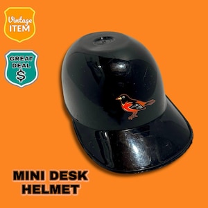 Op de afbeelding: Een zwarte mini-bureauhelm met een glanzende afwerking, met een klein vogel-embleem in rood, zwart en oranje. De helm heeft een vizier en de tekst "MINI DESK HELMET" is zichtbaar. De achtergrond is oranje.