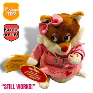 Peut inclure: Un renard en peluche portant un manteau rose avec un cœur sur la poitrine. Le renard a un nœud rose sur la tête et tient une pancarte qui dit "Try Me! Light Up I Sing and Dance I Got You Babe". Le renard est un jouet qui chante et danse.