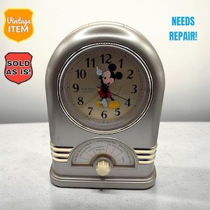 May include: A silver-toned Seiko quartz clock featuring a Mickey Mouse illustration on the face with black numerals. The base includes a dial and text listing song titles. The clock displays the text VINTAGE ITEM, SOLD AS IS, and NEEDS REPAIR.
