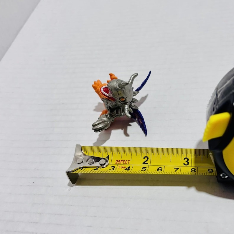 Puede incluir: Una peque&ntilde;a figura de criatura naranja y plateada con detalles en azul, que presenta una pieza met&aacute;lica en la cabeza y garras afiladas. Una cinta m&eacute;trica amarilla est&aacute; colocada junto a la figura, indicando una longitud de unos 3,8 cm.