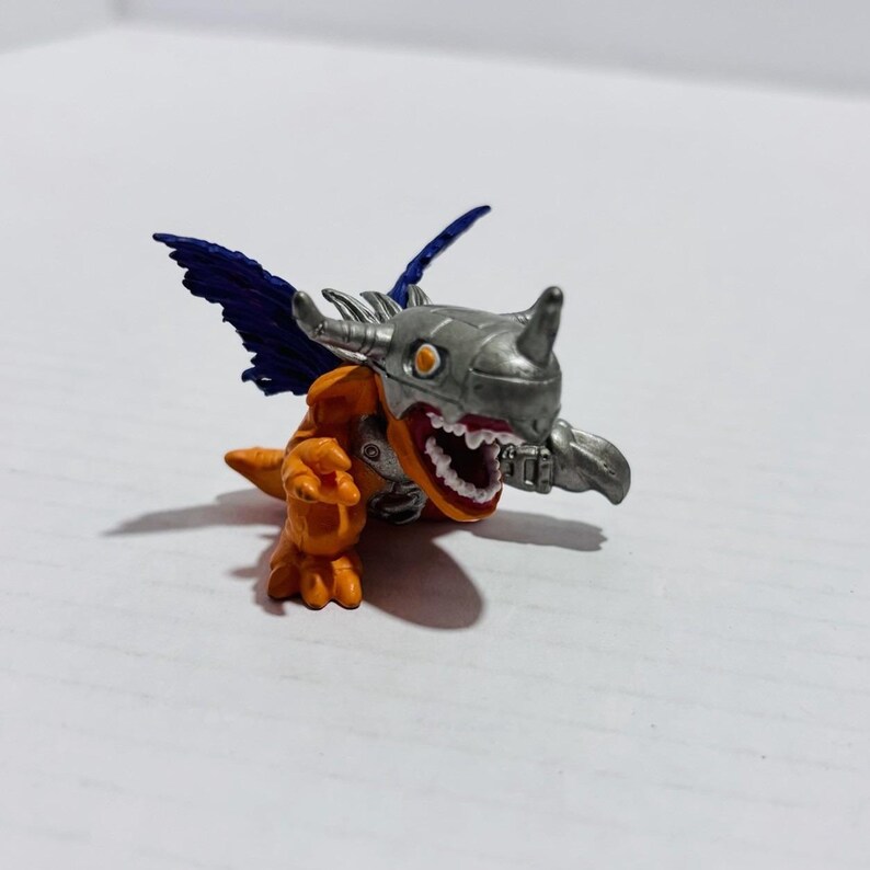 Puede incluir: Una peque&ntilde;a figura de criatura naranja con un casco met&aacute;lico plateado que presenta un solo cuerno. La figura tiene alas azul oscuro, dientes blancos afilados y la boca abierta. Est&aacute; en una postura din&aacute;mica con una garra extendida.