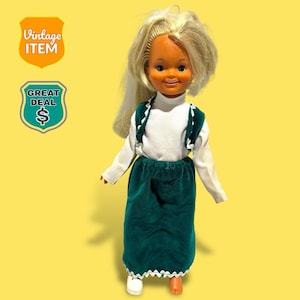 May include: A vintage doll with blonde hair, dressed in a white long-sleeved top, a green velvet skirt, and a green vest with white trim. The doll has a smiling face and is set against a yellow backdrop. The image includes "Vintage Item" and "Great Deal" badges.