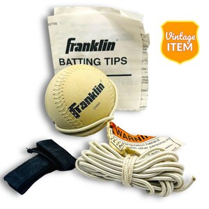 Op de afbeelding: Een witte honkbal met een zwart Franklin-logo en een wit touw eraan vastgemaakt. De honkbal ligt op een wit oppervlak met een witte tas met de tekst "Franklin Batting Tips" erop. Er is een zwarte polsband en een waarschuwingslabel op het touw.