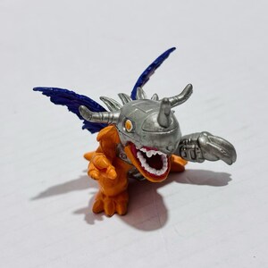 Puede incluir: Una peque&ntilde;a figura de criatura naranja con un casco met&aacute;lico plateado que presenta un solo cuerno. La figura tiene alas de color azul oscuro, una boca muy abierta con dientes blancos visibles y una mano con garras plateadas extendida.