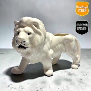 以下が含まれることがあります： 光沢のある仕上げが施されたクリーム色の陶器製ライオン型プランター。ライオンは立った姿勢で、詳細なタテガミと鼻と口に黒い塗装のアクセントが施されています。ライオンの背中の上部には開いた空洞があります。