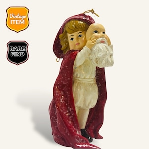 May include: A vintage Christmas bauble depicting a child wearing a red hooded cloak holding a Santa Claus mask. The bauble is made of white and red painted resin and has a glittery finish.