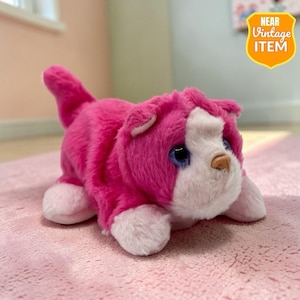 Op de afbeelding: Een roze pluche kattenknuffel met witte accenten op het gezicht en de poten. Het speelgoed heeft blauwe ogen en een bruine neus. Het label luidt "NEAR VINTAGE ITEM". De kat ligt op een roze ondergrond.