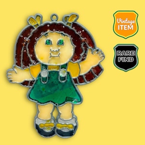 May include: A stained glass doll pendant with a yellow face, green eyes, and brown hair. The doll wears a green dress and yellow shirt. The pendant has a silver border and a loop for hanging. Two shield-shaped labels read "Vintage Item" and "Rare Find."
