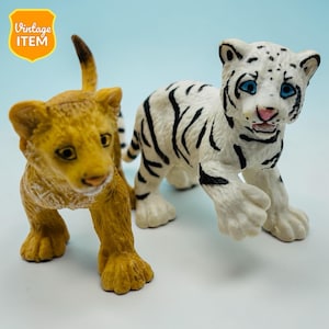 Könnte beinhalten: Zwei Spielzeugfiguren: ein goldfarbenes Löwenjunges und ein weißes Tigerjunges mit schwarzen Streifen. Das Löwenjunge hat einen erhobenen Schwanz und blickt nach links. Das Tigerjunge hat blaue Augen und eine rosa Nase. Ein Vintage-Artikel-Abzeichen befindet sich in der oberen linken Ecke.
