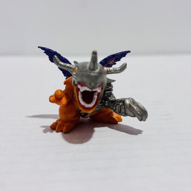 Puede incluir: Una peque&ntilde;a figura de criatura naranja y plateada con un casco met&aacute;lico, dientes afilados y alas azul oscuro. La figura tiene la boca muy abierta, un cuerno en la cabeza y una mano con garras extendida hacia adelante en una pose din&aacute;mica.