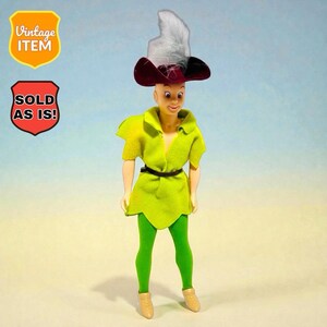 May include: A vintage doll wearing a green tunic, green leggings, and tan shoes. The doll has a burgundy and white feathered hat. The text "Vintage Item" and "Sold As Is!" are visible.
