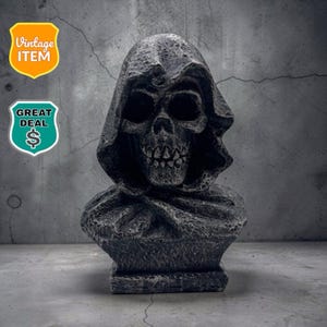 May include: A gray, textured stone-like bust of a skull wearing a hooded cloak. The sculpture features deep, hollow eye sockets, a detailed nasal cavity, and a row of teeth. The surface has a weathered, rugged appearance with dark shading.