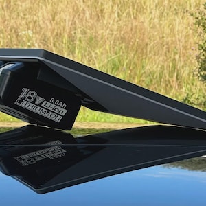 Può includere: Un pacco batteria nero agli ioni di litio da 18 V con capacità di 8,0 Ah, collegato a un dispositivo nero angolato. Il dispositivo è appoggiato su una superficie riflettente, con uno sfondo naturale di erba e fogliame.