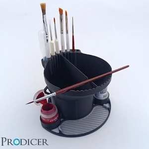 May include: A black paint brush holder with multiple compartments, holding various paint brushes of different sizes and shapes. A red paint pot and a long paint brush are also visible. The holder has a gray, textured base.