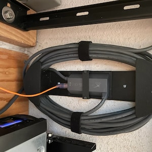 Könnte beinhalten: Ein schwarzes Kabelmanagementsystem mit einem grauen Kabel, das um es herumgewickelt ist. Ein schwarzes Gerät mit einem weißen Etikett ist an dem System montiert. Das System ist an einer Wand befestigt.