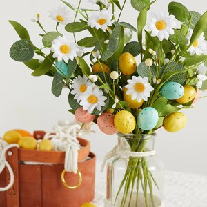 May include: A white vase with a rope design around the middle, filled with a bouquet of white daisies, green leaves, and colourful speckled Easter eggs.