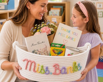 May include: A white woven rope basket featuring the name Miss Banks embroidered in colourful yarn on the front. The basket contains a bouquet of flowers, a book labelled Miss Banks, a box of Crayola crayons, and a card reading Thank You Miss Banks.
