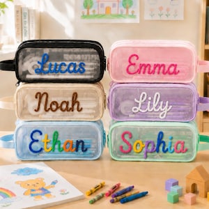May include: Six rectangular, transparent pencil cases with side handles, each featuring a personalized name in raised, colorful yarn lettering. The cases are stacked in two columns and come in black, beige, light blue, pink, purple, and mint green.