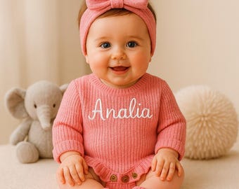May include: A baby wearing a pink ribbed knit romper with the name "Analia" embroidered in white. The outfit includes a matching headband and socks. A gray stuffed elephant and a white textured ball are in the background.