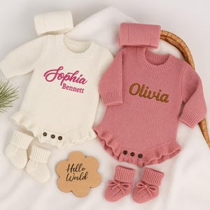 Könnte beinhalten: Zwei gestrickte Baby-Strampler, einer cremefarben und einer rosa, jeweils mit passendem Stirnband und Socken. Der cremefarbene Strampler hat "Sophia Bennett" in Rosa gestickt, und der rosa Strampler hat "Olivia" in Gold gestickt. Ein Holzschild sagt "Hello World".
