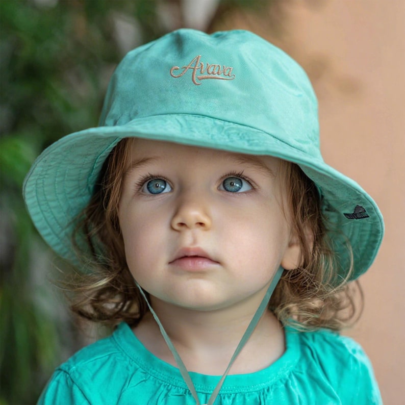 Pu&ograve; includere: Un cappello a secchiello turchese chiaro con la parola "Arava" ricamata in corsivo. Il cappello ha un'ampia tesa e un sottogola. Il bambino indossa una maglietta turchese abbinata. Il cappello ha un piccolo logo sul lato.