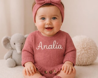 May include: A baby wearing a matching rose-colored outfit, including a headband with a bow, a long-sleeved romper with the name "Analia" embroidered in white, and leg warmers. A plush elephant toy is visible in the background.
