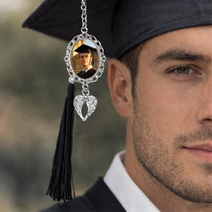 Pu&ograve; includere: Un nappa del cappello di laurea con un ciondolo fotografico argentato. Il ciondolo presenta una cornice ovale con una foto di un laureato, un ciondolo a forma di cuore con ali d'angelo e una lunga nappa nera. Il cappello &egrave; nero.