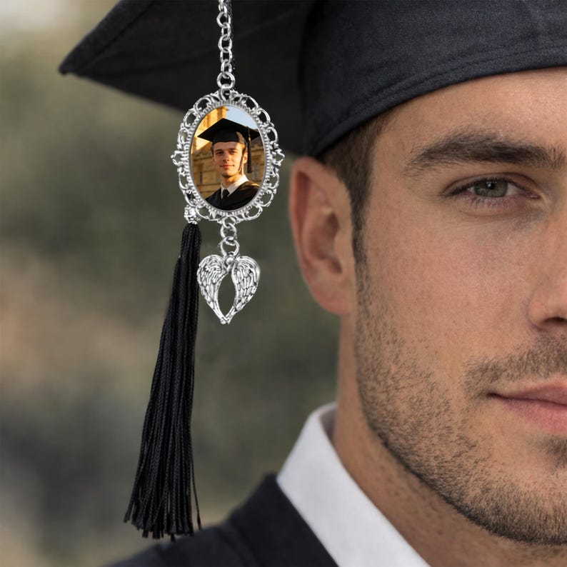 Pu&ograve; includere: Un nappa del cappello di laurea con un ciondolo fotografico argentato. Il ciondolo presenta una cornice ovale con una foto di un laureato, un ciondolo a forma di cuore con ali d'angelo e una lunga nappa nera. Il cappello &egrave; nero.