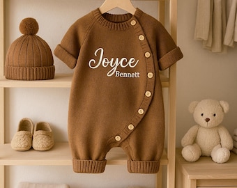 May include: A brown knitted baby romper with the name "Joyce Bennett" in white script. The romper has short sleeves and is buttoned on the side. A matching brown knitted hat with a pom-pom, a pair of baby shoes, and a teddy bear are also visible.