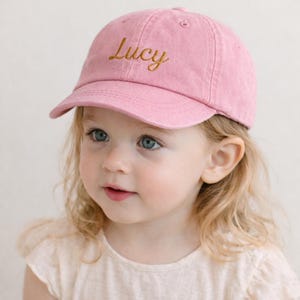 Pode incluir: Boné de beisebol rosa com o nome "Lucy" bordado em fio dourado. O boné tem uma aba curva e um design clássico de seis painéis. O chapéu é feito de um tecido macio e texturizado.