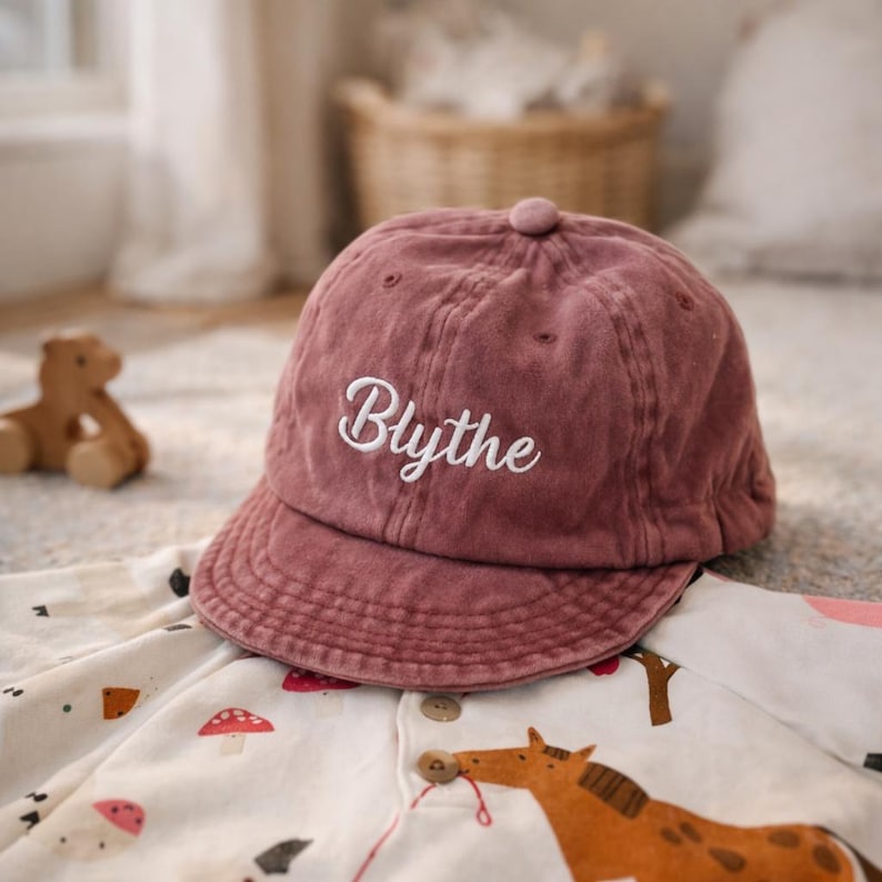 K&ouml;nnte beinhalten: Staubrosa Baseballkappe mit dem Namen "Blythe" in wei&szlig;er Schrift gestickt. Die Kappe besteht aus einem weichen, strukturierten Stoff und hat einen gebogenen Schirm. Die Kappe liegt auf einem bedruckten Strampler.